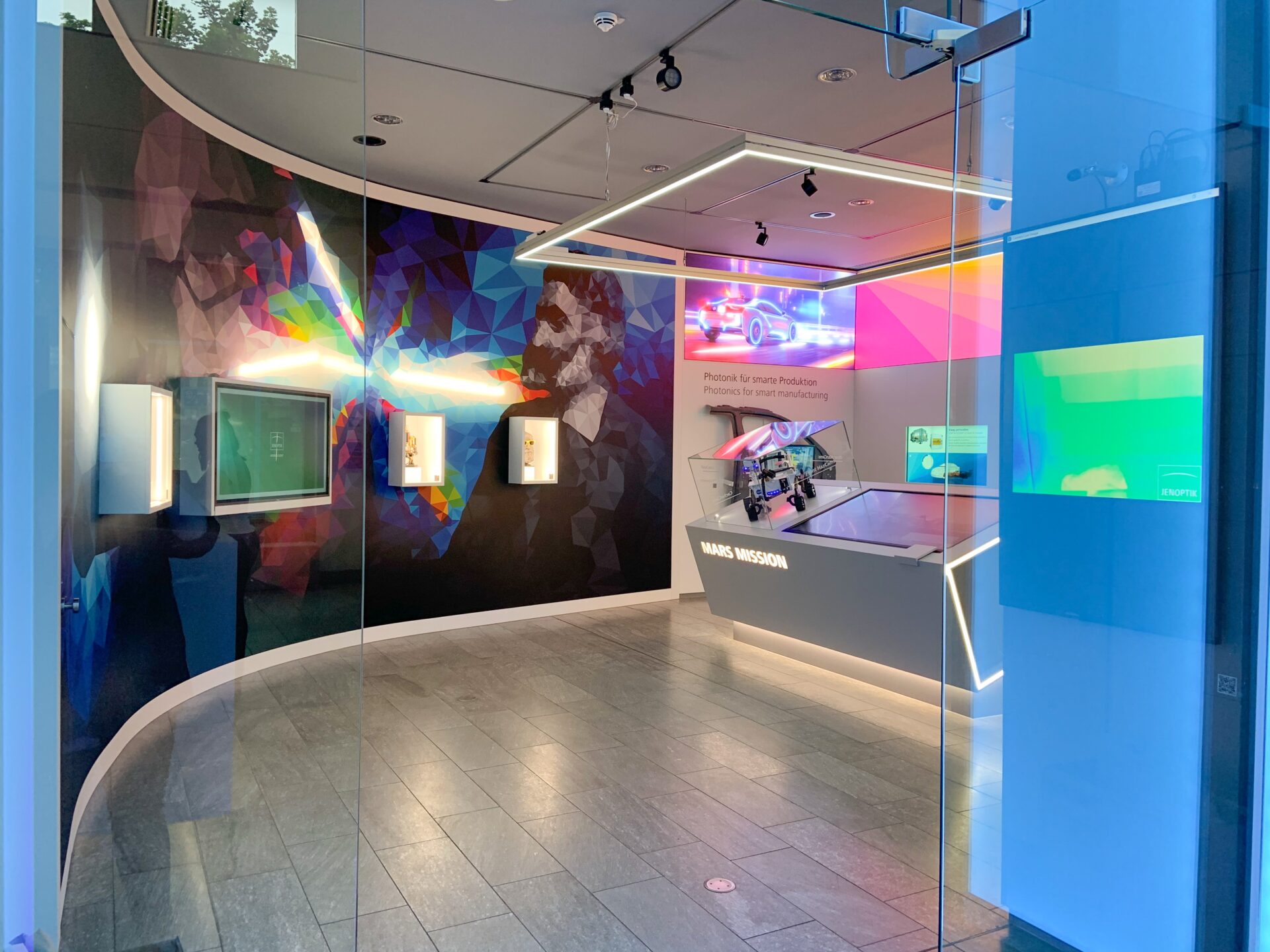 Ausstellungsbereich der JENOPTIK AG mit beleuchteten Vitrinen, Produkten und einem bunten Wandgemälde mit dem abstrahierten Portrait von Carl Zeiss