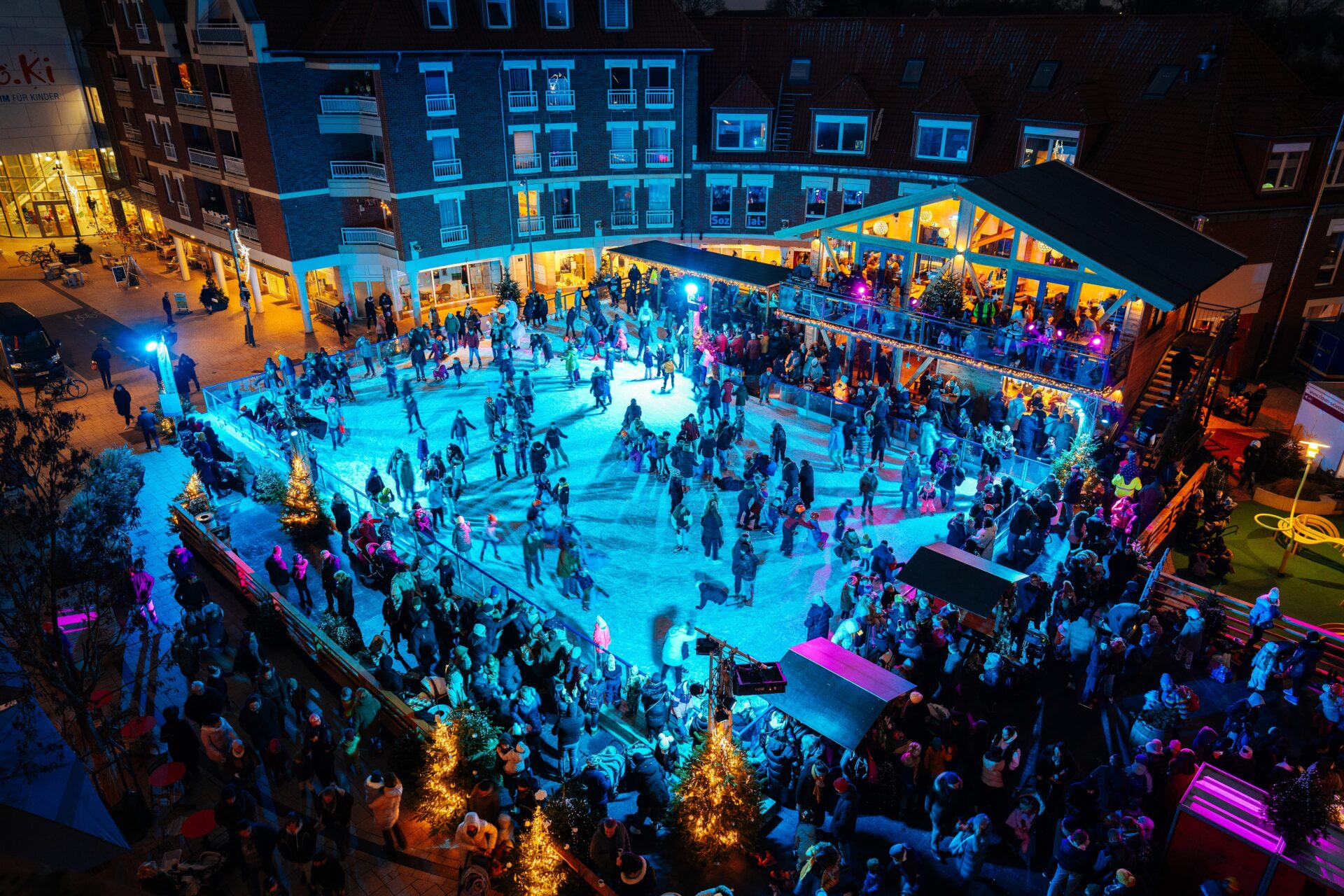 Blick auf eine belebte Eislaufbahn bei Nacht, umgeben von festlich beleuchteten Gebäuden und Bäumen.