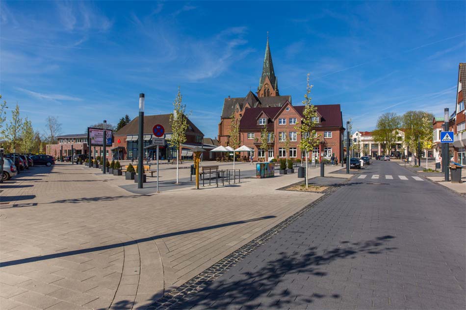 Marktplatz in Sögel mit Kirche im Hintergrund, Sitzbereich und Bäumen unter blauem Himmel
